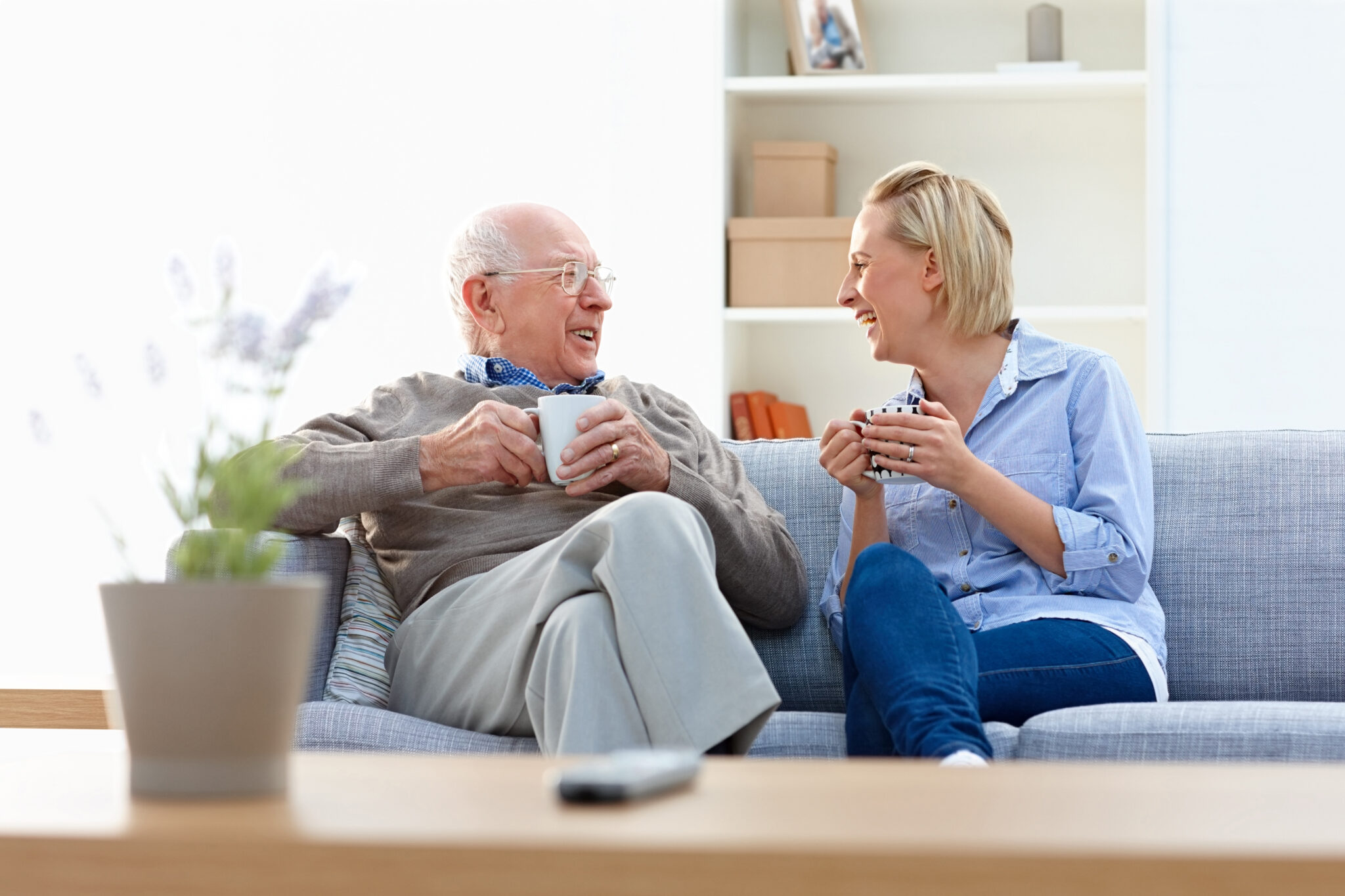Senior man and female carer sitting together on sofa enjoying conversation with coffee in living room - Indoors